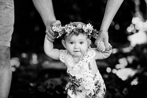Baby girl taking early steps while holding her parents' hands during an outdoor portrait session