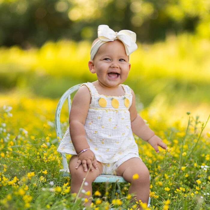 Smiling one year old girl sitting among yellow flowers during an outdoor milestone session
