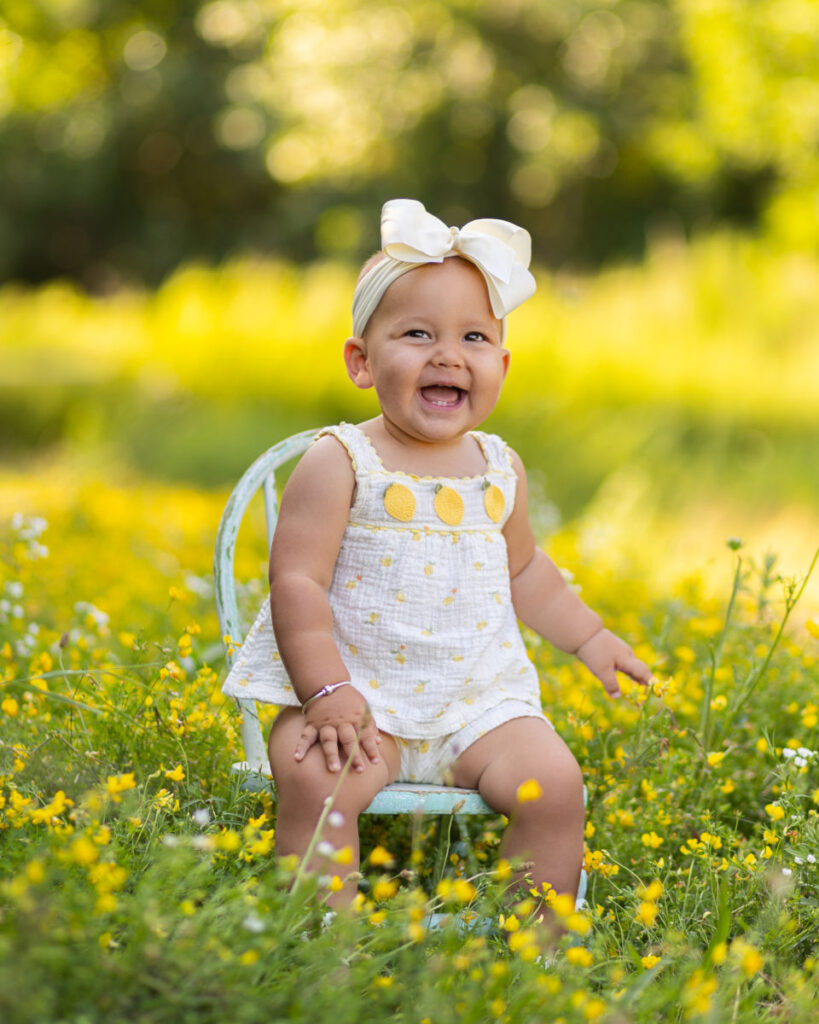 Smiling one year old girl sitting among yellow flowers during an outdoor milestone session