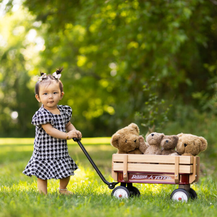 Toddler girl standing beside a wagon filled with teddy bears during an outdoor portrait session