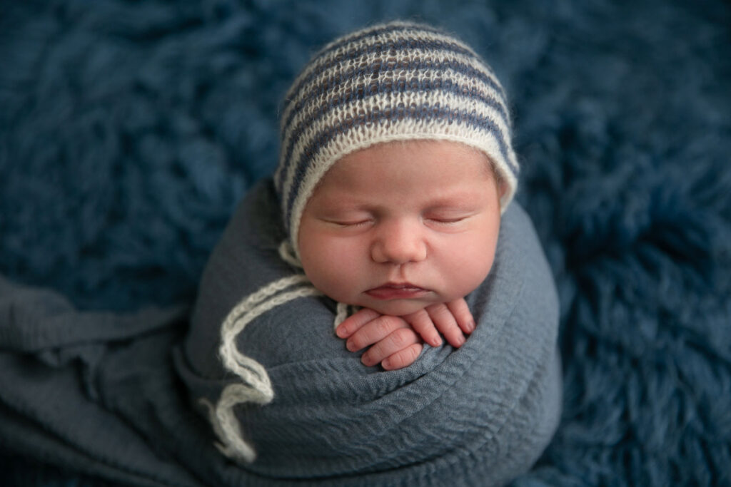 Sleeping newborn wrapped in blue fabric wearing a striped knit bonnet during a newborn photography session