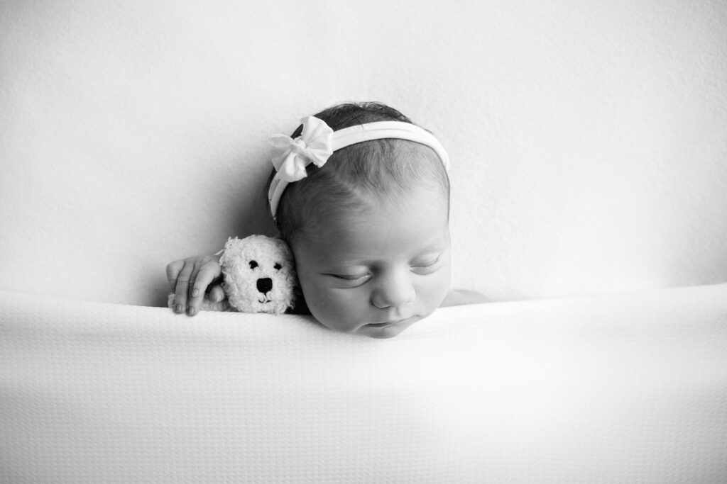 Sleeping newborn baby beside small teddy bear during black-and-white studio portrait