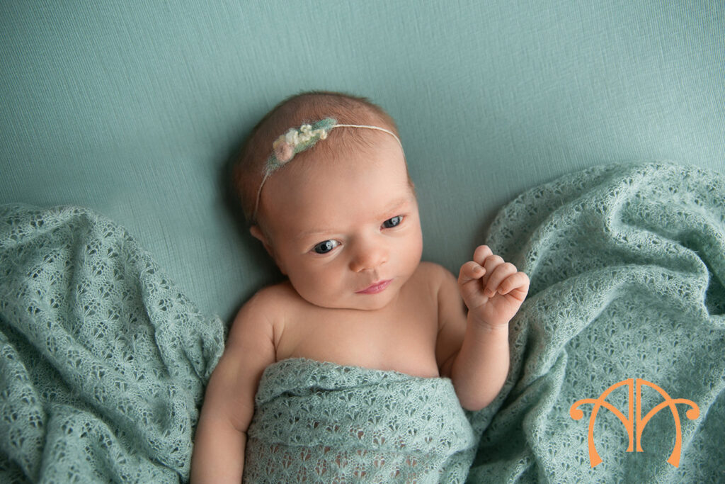 Awake newborn baby girl wrapped in mint fabric looking toward camera during studio portrait session