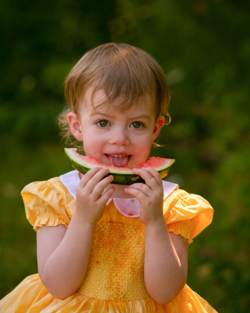 Toddler smiling while eating watermelon during summer portrait session.