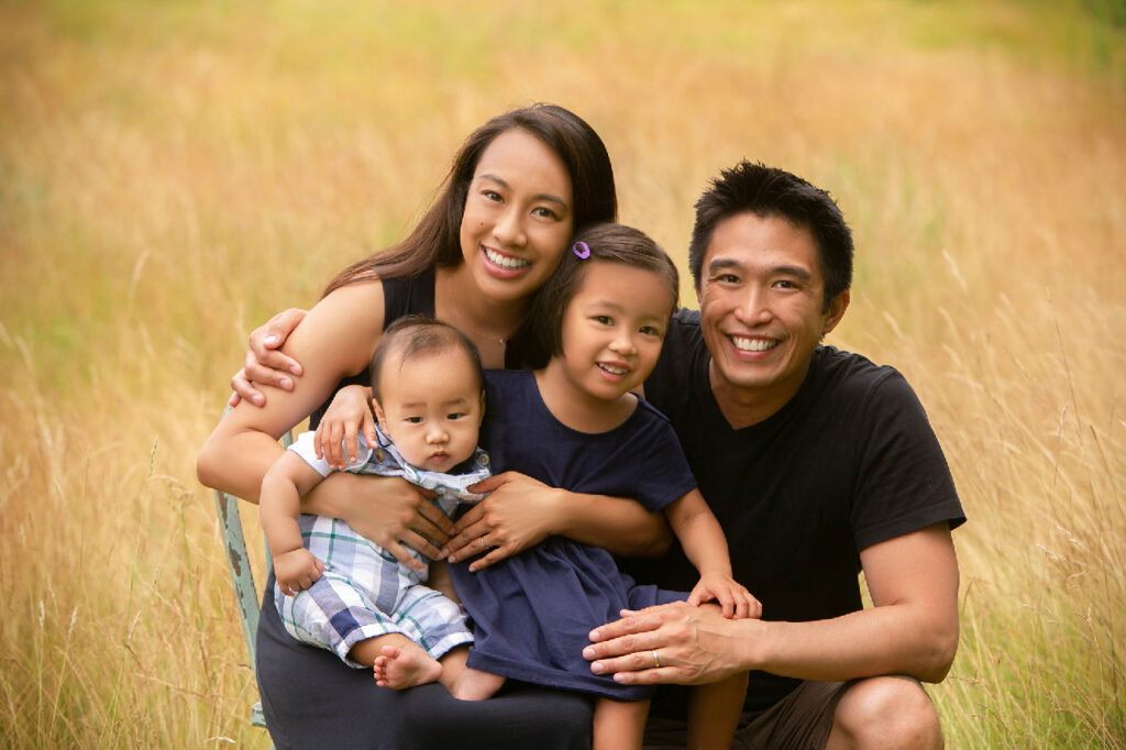 Family of four smiling together outdoors in grassy field during portrait session.