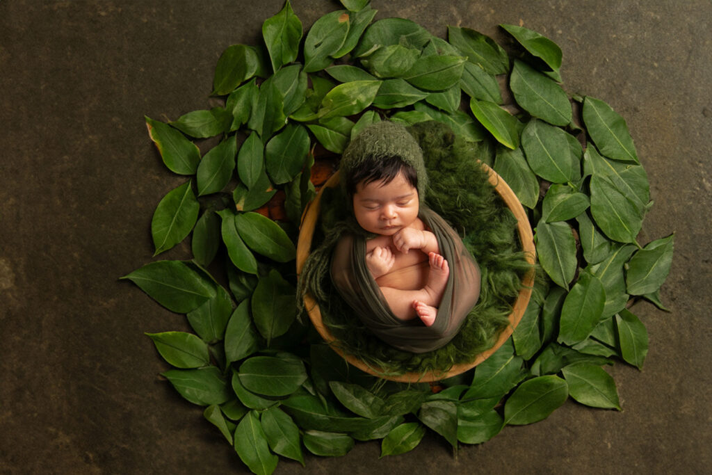 Sleeping newborn baby wrapped in soft green fabric surrounded by natural leaves in a studio portrait