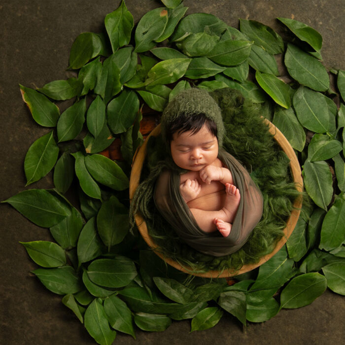 Sleeping newborn baby wrapped in soft green fabric surrounded by natural leaves in a studio portrait