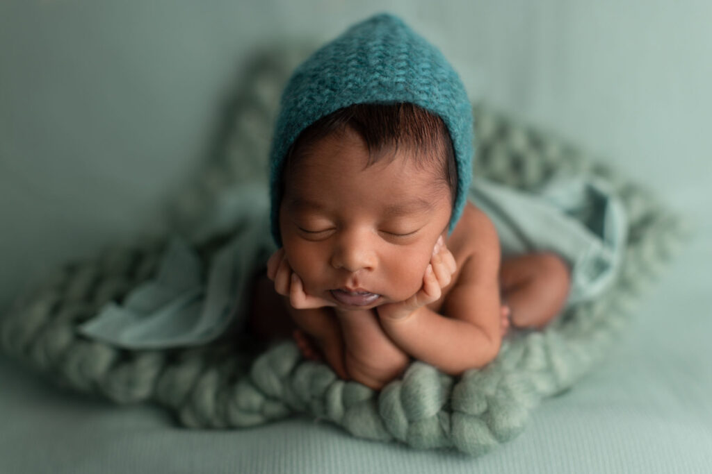 Sleeping newborn baby wearing teal bonnet posed on soft textured blanket during studio session