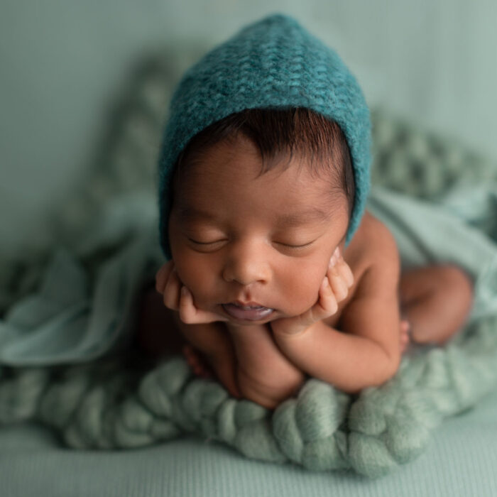 Sleeping newborn baby wearing teal bonnet posed on soft textured blanket during studio session