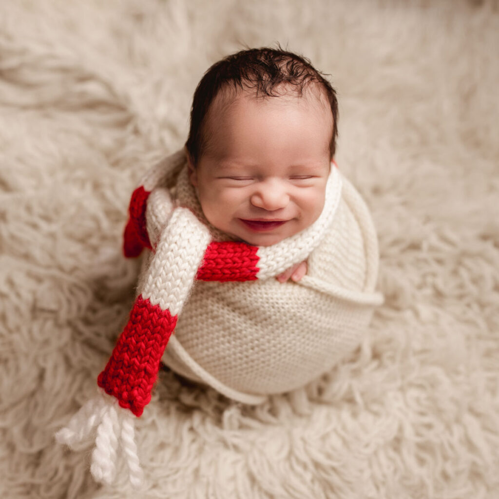 Smiling newborn baby boy wrapped in cream fabric with a holiday striped red knit scarf