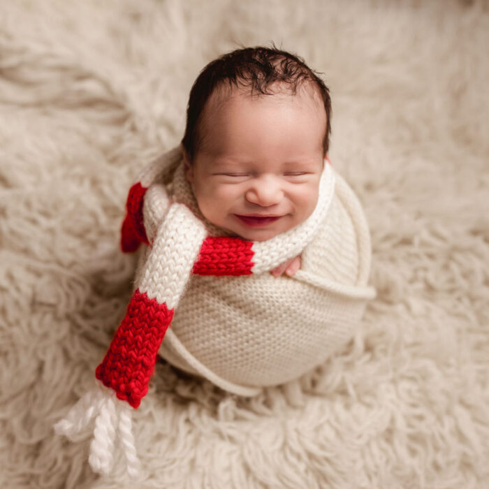 Smiling newborn baby boy wrapped in cream fabric with a holiday striped red knit scarf