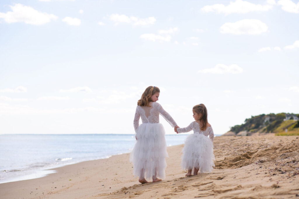 Two young sisters in white dresses walking together on a sandy beach during a family photography session