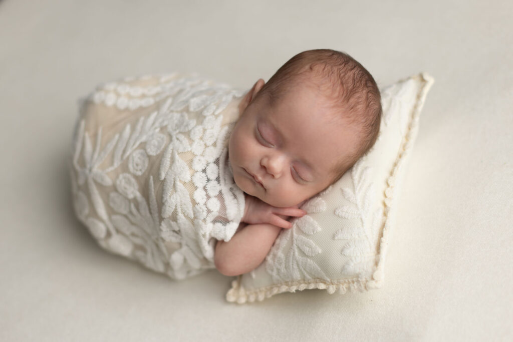 Sleeping newborn baby wrapped in cream fabric resting on decorative pillows
