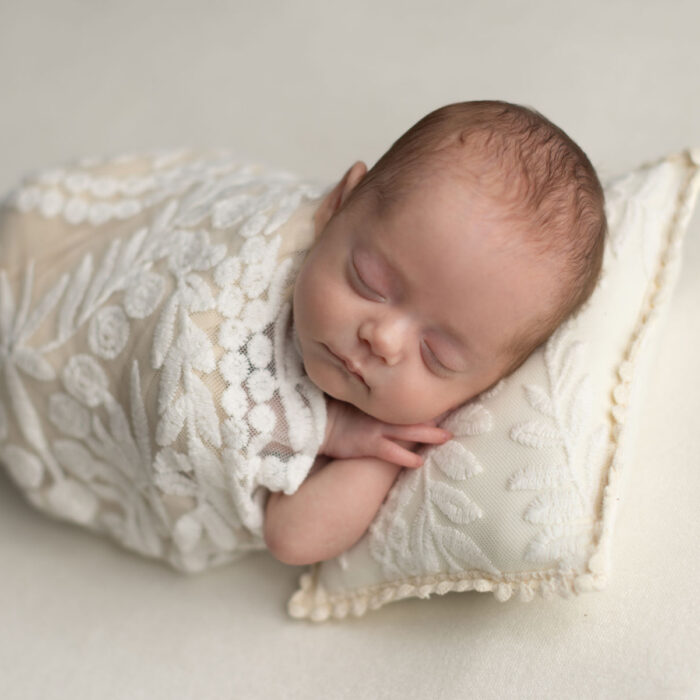 Sleeping newborn baby wrapped in cream fabric resting on decorative pillows