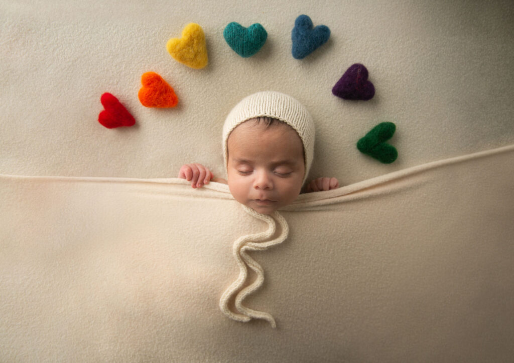 Sleeping newborn baby tucked beneath a blanket surrounded by colorful heart decorations