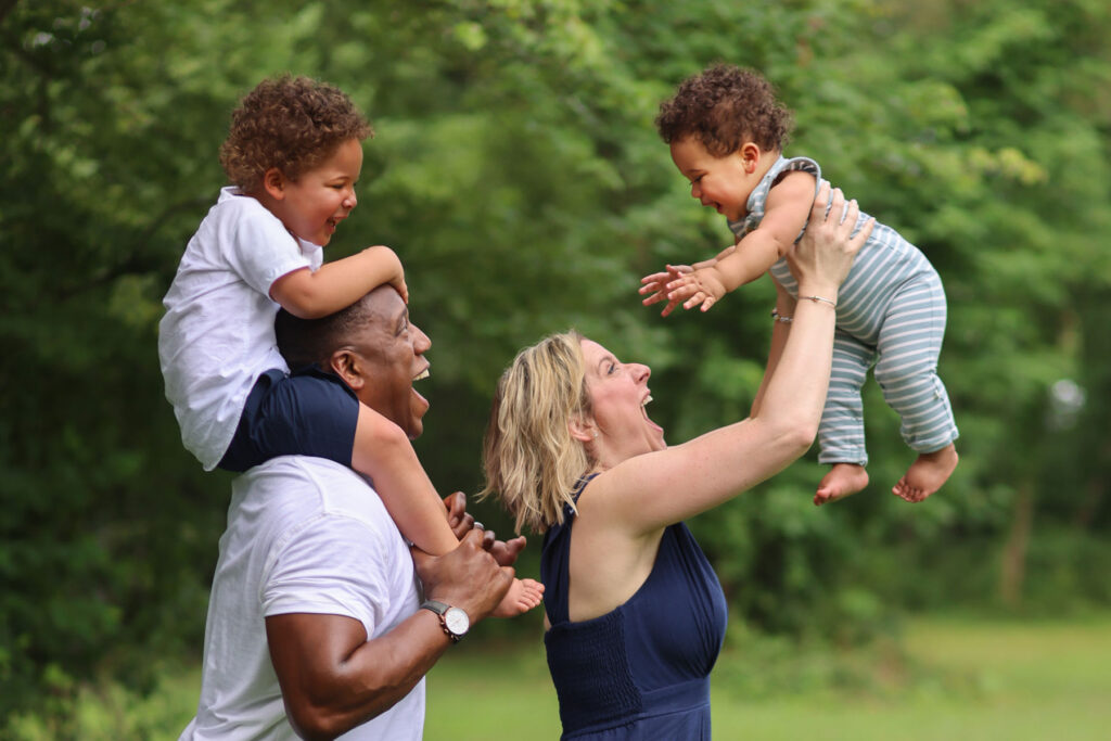 Family portrait outdoors with parents lifting children and sharing playful moments.