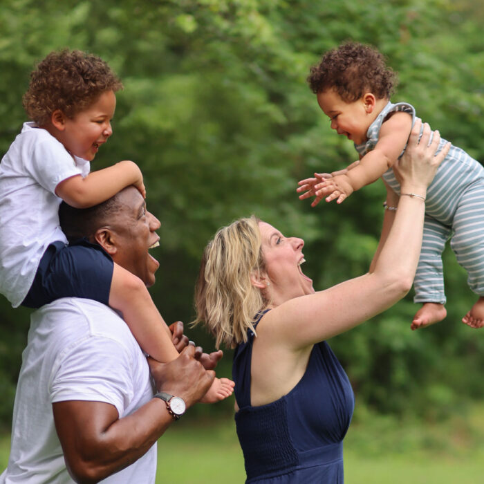 Family portrait outdoors with parents lifting children and sharing playful moments.
