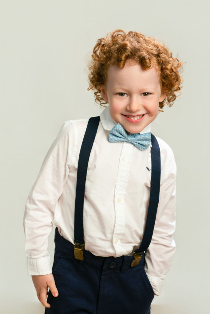 Young boy with curly reddish hair wearing suspenders and bow tie during portrait session.