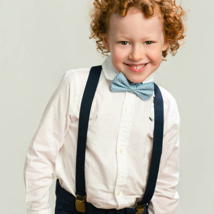 Young boy with curly reddish hair wearing suspenders and bow tie during portrait session.