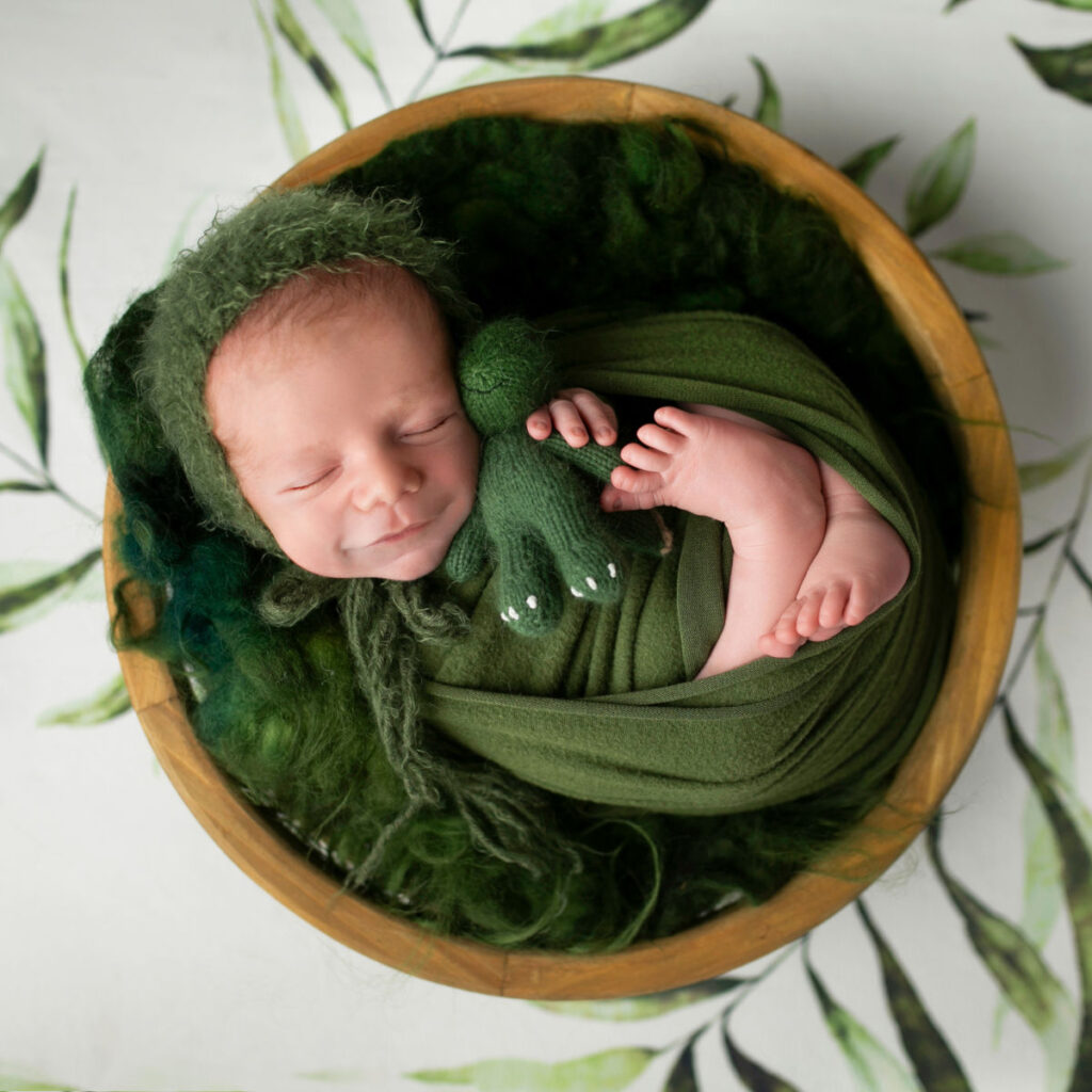 Sleeping newborn baby wrapped in green posing in a wooden bowl during studio newborn photography session