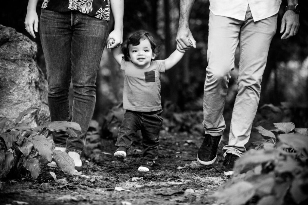 Toddler walking between parents during a black-and-white outdoor family portrait