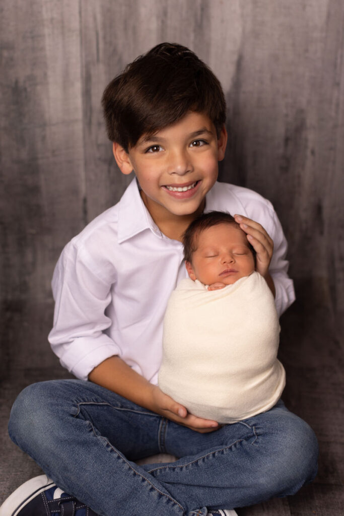Smiling young boy holding his swaddled newborn baby sister during a professional sibling newborn portrait session