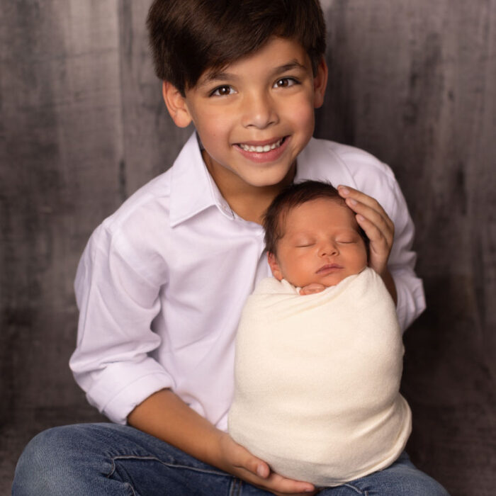 Smiling young boy holding his swaddled newborn baby sister during a professional sibling newborn portrait session
