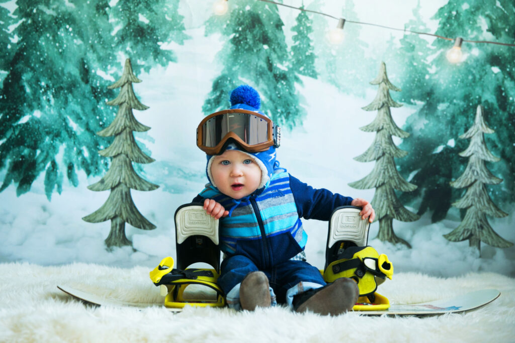Baby in winter clothing posing with snowboard equipment during a themed portrait session