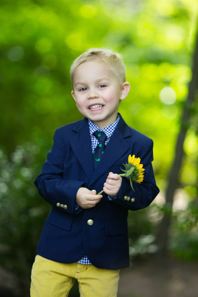Smiling three year old boy in a navy blazer holding a sunflower during an outdoor child portrait session