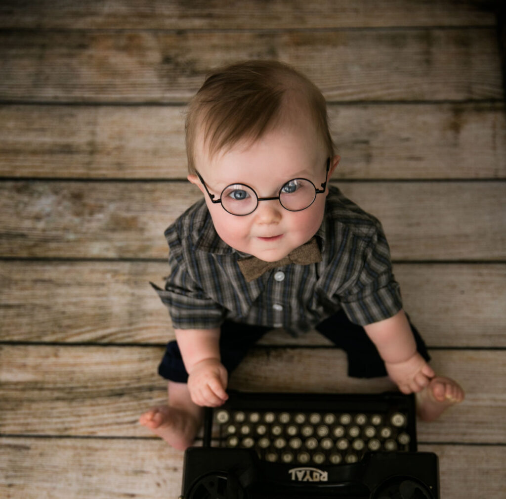 Baby boy wearing glasses sitting beside vintage typewriter during milestone sitter photography session