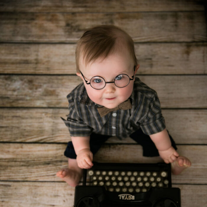 Baby boy wearing glasses sitting beside vintage typewriter during milestone sitter photography session