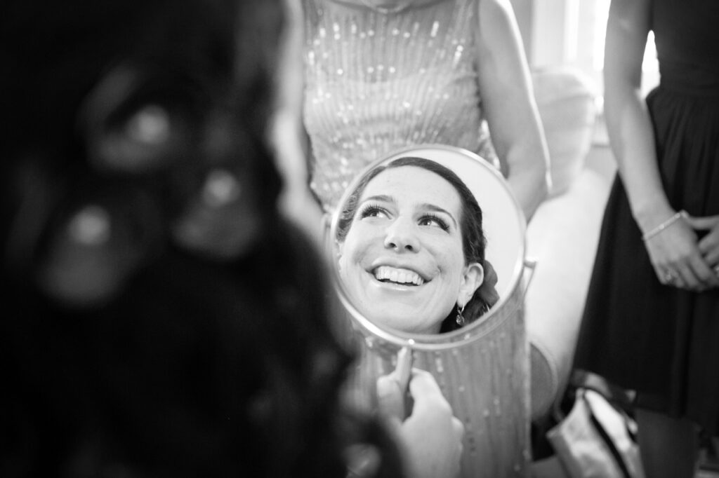 Bride smiling in a mirror while getting ready with her bridal party before the wedding ceremony
