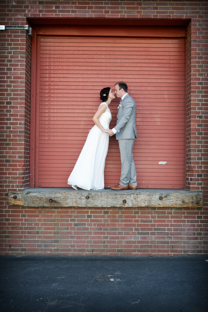Bride and groom kissing on a loading dock in front of a red industrial door during a creative wedding portrait session