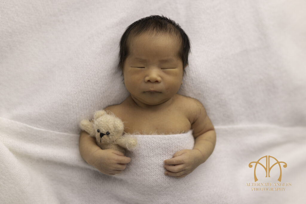 Japanese newborn baby wrapped in white blanket holding teddy bear in Boston studio