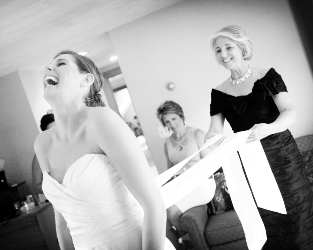 Bride laughing as her mother helps tie her wedding dress while getting ready before the ceremony