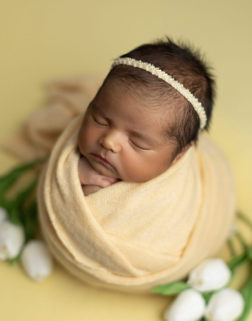 Sleeping newborn baby girl wrapped in soft yellow fabric with white tulips during a newborn photography session in Norwood, MA