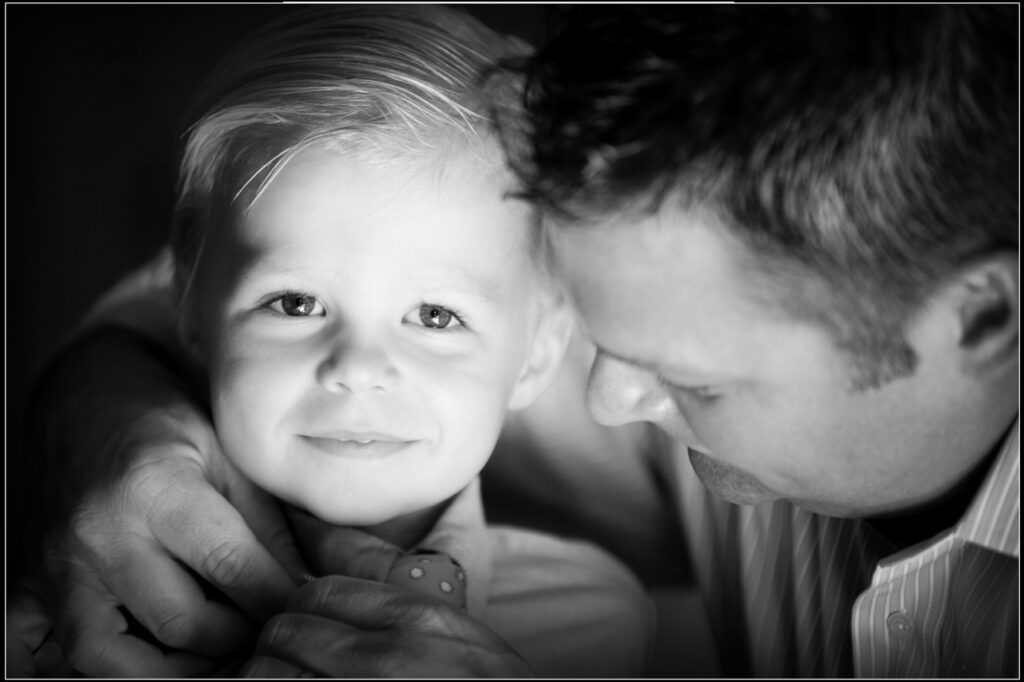 Young ring bearer smiling while his dad helps tie his tie before a wedding ceremony