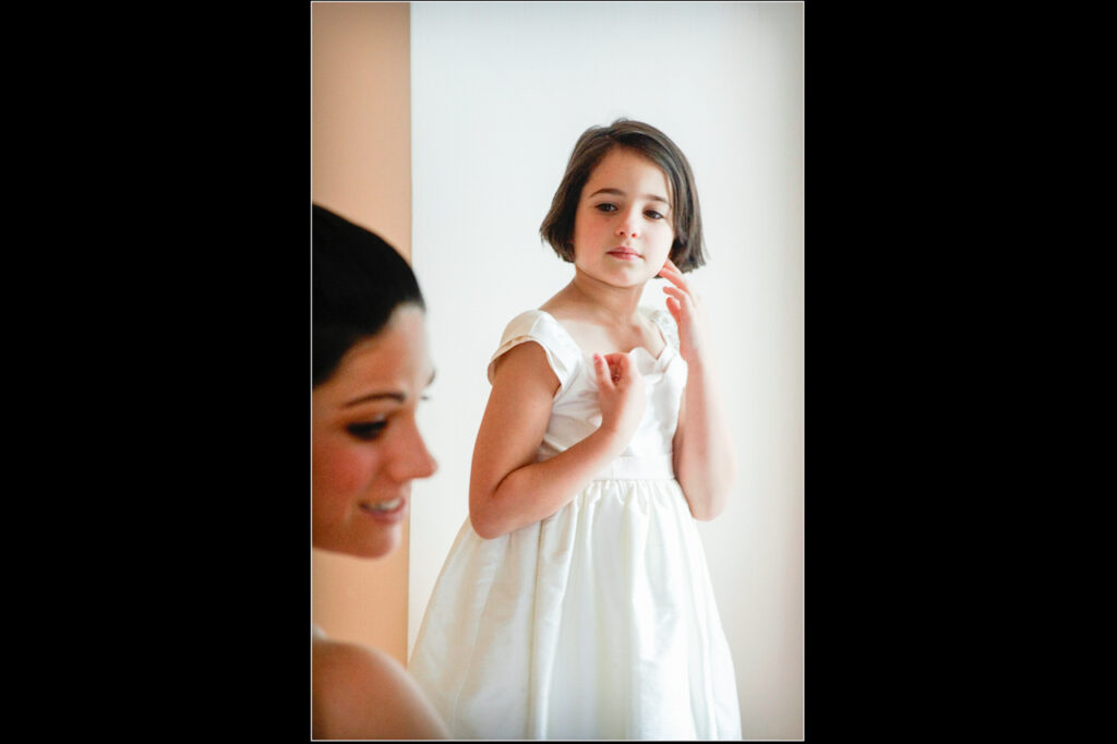 Young flower girl in a white dress standing near the bride during a soft natural-light wedding portrait