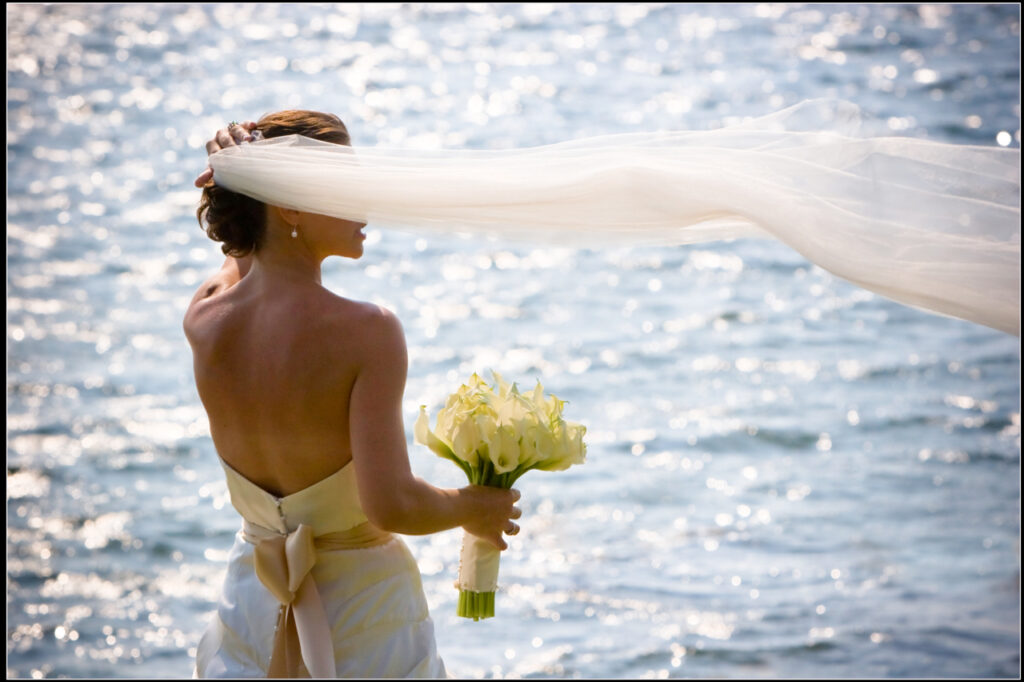 Bride holding white bouquet of lilies with veil blowing in the wind during a waterfront wedding portrait in Marion, Massachusetts